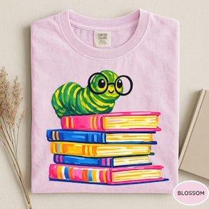 Pu&ograve; includere: Una t-shirt rosa chiaro con la grafica di un bruco a strisce verdi e gialle che indossa occhiali tondi neri, appoggiato su una pila di cinque libri colorati. La maglietta presenta un'etichetta del marchio Comfort Colors stampata sul collo.