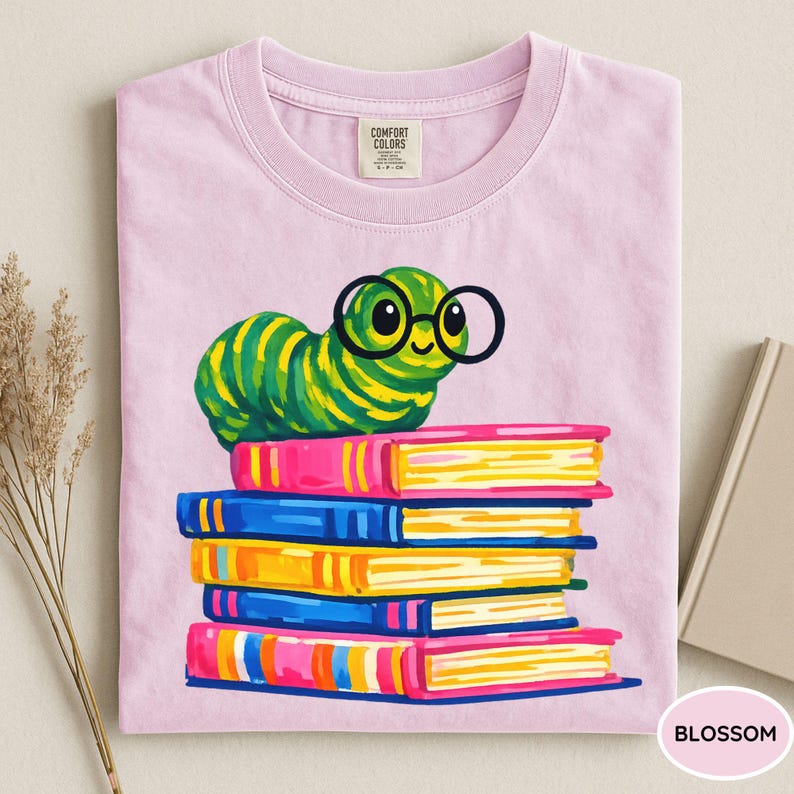Pu&ograve; includere: Una t-shirt rosa chiaro con la grafica di un bruco a strisce verdi e gialle che indossa occhiali tondi neri, appoggiato su una pila di cinque libri colorati. La maglietta presenta un'etichetta del marchio Comfort Colors stampata sul collo.