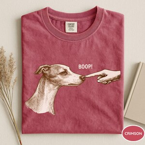 May include: A crimson crew neck t-shirt featuring a brown graphic illustration of a dog being touched on the nose by a finger with the text BOOP! printed above. The shirt has a Comfort Colors brand label on the inside of the collar.