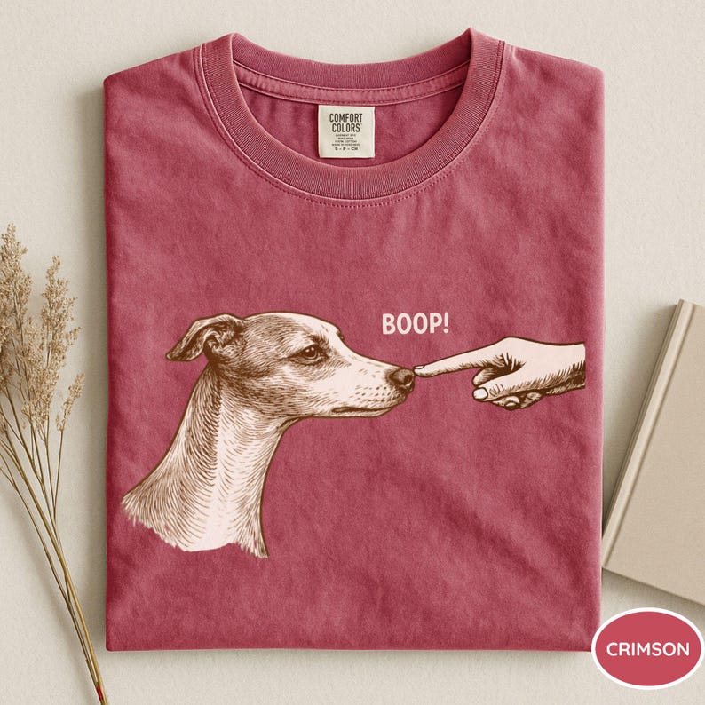 May include: A crimson crew neck t-shirt featuring a brown graphic illustration of a dog being touched on the nose by a finger with the text BOOP! printed above. The shirt has a Comfort Colors brand label on the inside of the collar.