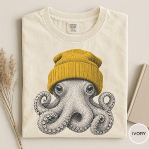 May include: Ivory-colored t-shirt featuring a detailed illustration of an octopus wearing a mustard yellow beanie. The octopus has large, expressive eyes and detailed tentacles. The shirt is made of Comfort Colors material.