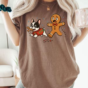 May include: Espresso-colored t-shirt featuring a cartoon French Bulldog wearing a Christmas sweater, holding a gingerbread man, and a scared gingerbread man. The shirt has a crew neck and short sleeves. The word "Espresso" is printed on the bottom right.
