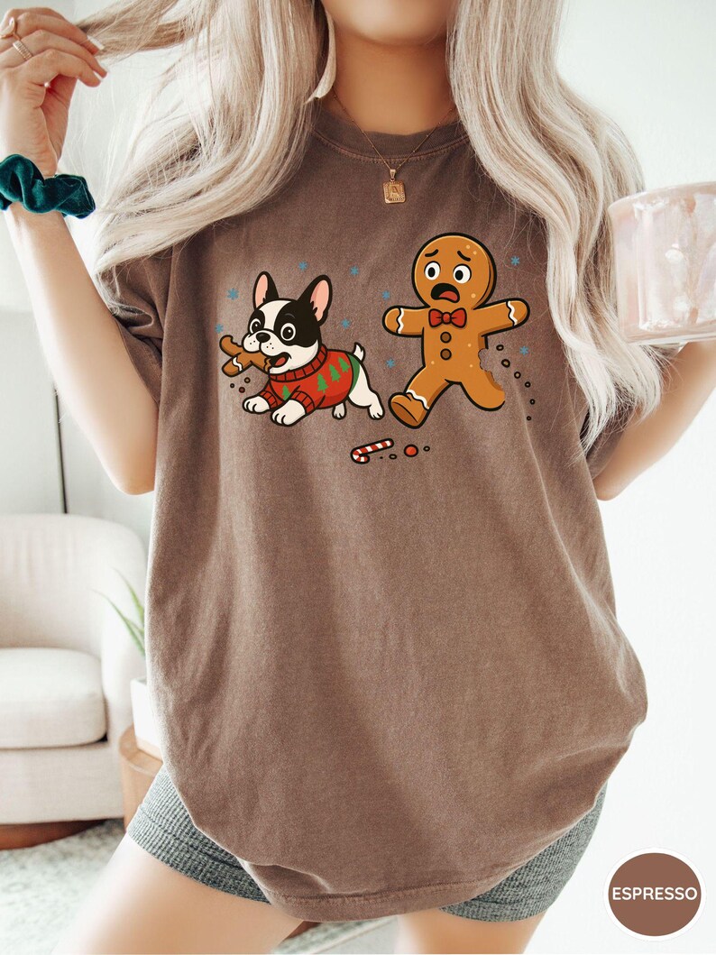 May include: Espresso-colored t-shirt featuring a cartoon French Bulldog wearing a Christmas sweater, holding a gingerbread man, and a scared gingerbread man. The shirt has a crew neck and short sleeves. The word "Espresso" is printed on the bottom right.