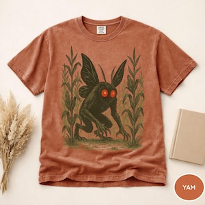 K&ouml;nnte beinhalten: Rostfarbenes T-Shirt mit einer dunkelgr&uuml;nen Kreatur mit roten Augen und gro&szlig;en Fl&uuml;geln, die in einem Maisfeld steht. Die Grafik ist auf einem Comfort Colors-T-Shirt gedruckt. Ein Buch und Trockenblumen sind im Hintergrund. Das Wort "YAM" steht auf einem kleinen orangefarbenen Kreis.