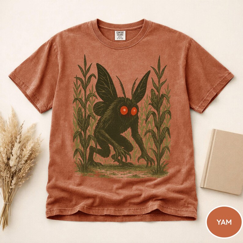 K&ouml;nnte beinhalten: Rostfarbenes T-Shirt mit einer dunkelgr&uuml;nen Kreatur mit roten Augen und gro&szlig;en Fl&uuml;geln, die in einem Maisfeld steht. Die Grafik ist auf einem Comfort Colors-T-Shirt gedruckt. Ein Buch und Trockenblumen sind im Hintergrund. Das Wort "YAM" steht auf einem kleinen orangefarbenen Kreis.