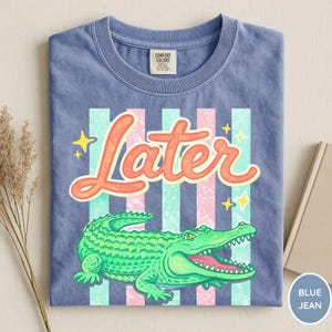 May include: Blue jean t-shirt with a graphic of a green alligator and the word "Later" in orange script. The design includes pink and teal stripes and yellow stars. The shirt is folded.