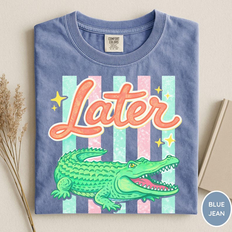 May include: Blue jean t-shirt with a graphic of a green alligator and the word "Later" in orange script. The design includes pink and teal stripes and yellow stars. The shirt is folded.
