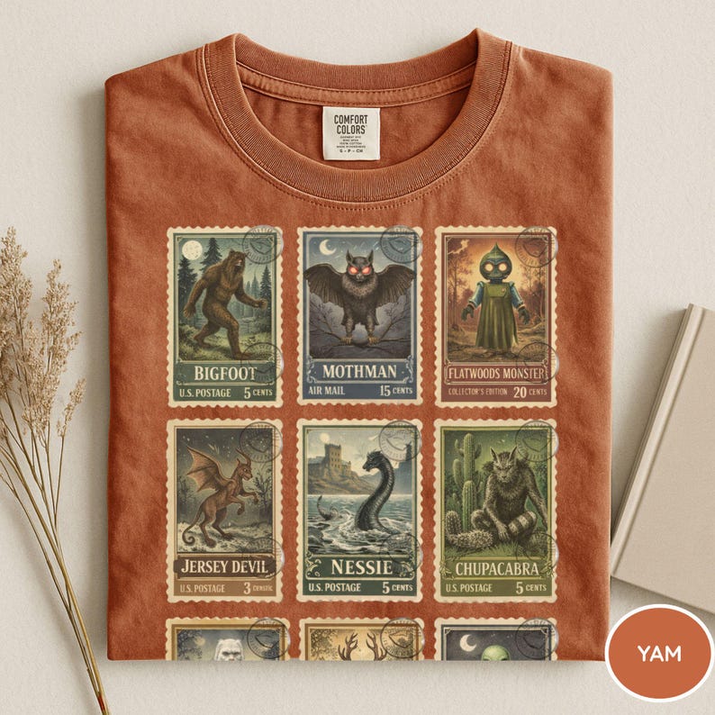 May include: A rust-colored t-shirt featuring a collection of vintage-style postage stamps. The stamps depict various cryptids, including Bigfoot, Mothman, and the Jersey Devil. The shirt has a crew neck and a tag that says "COMFORT COLORS."