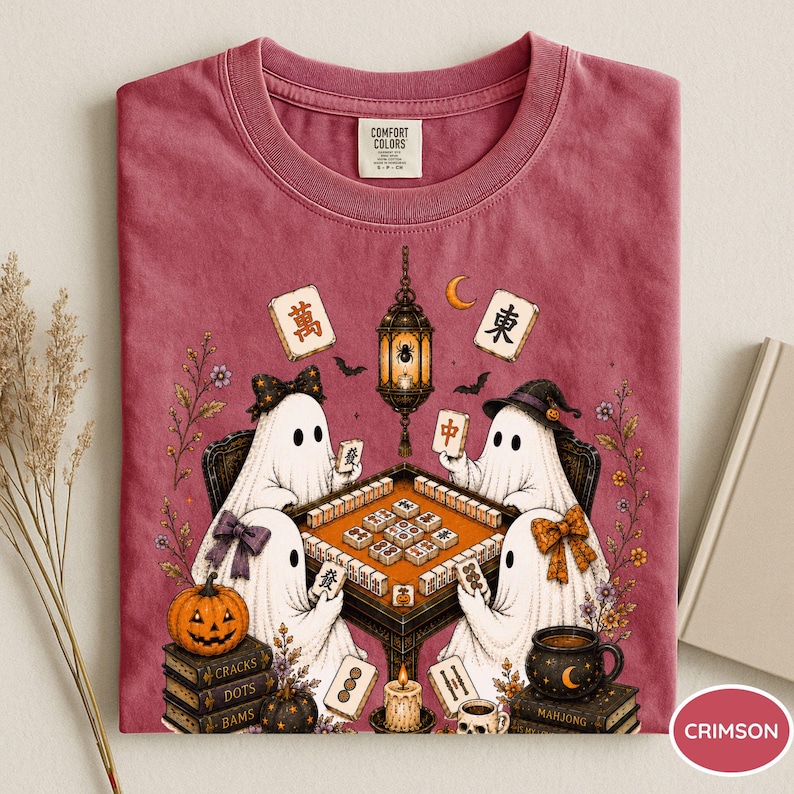 May include: A crimson t-shirt featuring a graphic of four white ghosts playing mahjong at a table. The design includes a hanging lantern, a jack-o'-lantern, stacks of books labeled Cracks, Dots, Bams, and Mahjong, and various mahjong tiles.