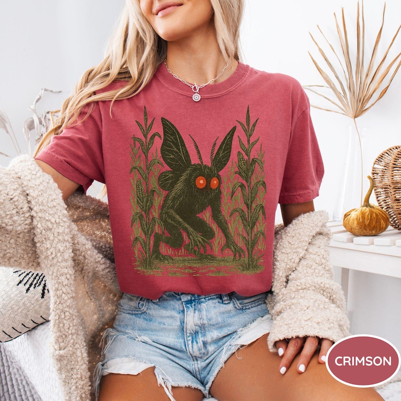 K&ouml;nnte beinhalten: Karmesinrotes T-Shirt mit einem gr&uuml;nen Kreaturen-Design, das gro&szlig;e Fl&uuml;gel und orangefarbene Augen aufweist, umgeben von Maisstauden. Das Shirt hat eine lockere Passform und kurze &Auml;rmel. Das Wort "CRIMSON" befindet sich in einem Kreis unten.