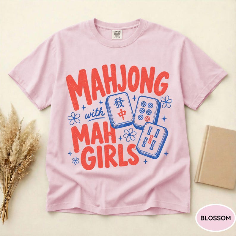 May include: A light pink short-sleeve t-shirt featuring red text that reads "MAHJONG with MAH GIRLS" and blue illustrations of three mahjong tiles decorated with small blue flowers and star accents across the center of the chest.