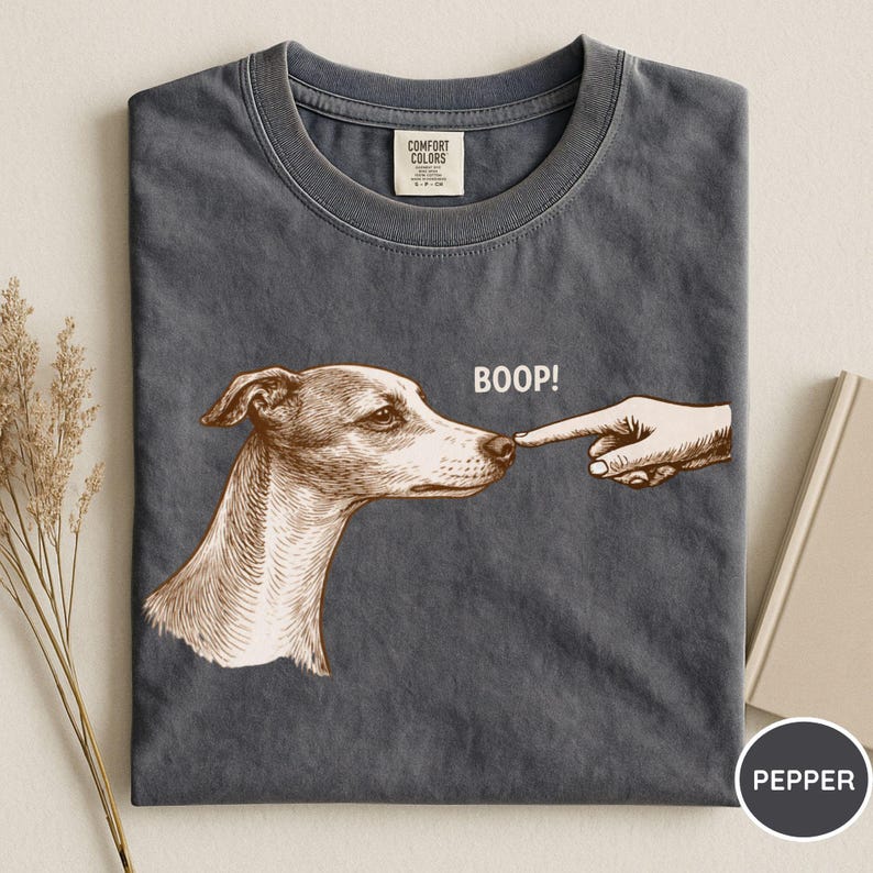 May include: A charcoal gray t-shirt featuring a brown ink-style illustration of a dog's head being touched on the nose by a human finger. The text BOOP! is printed in white above the dog's nose. The shirt has a Comfort Colors brand label.