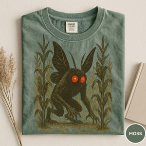 K&ouml;nnte beinhalten: Moosgr&uuml;nes T-Shirt mit einem schwarzen Wesen mit roten Augen und Fl&uuml;geln, umgeben von Maisstauden. Das T-Shirt hat einen Rundhalsausschnitt und ein Comfort Colors-Etikett.