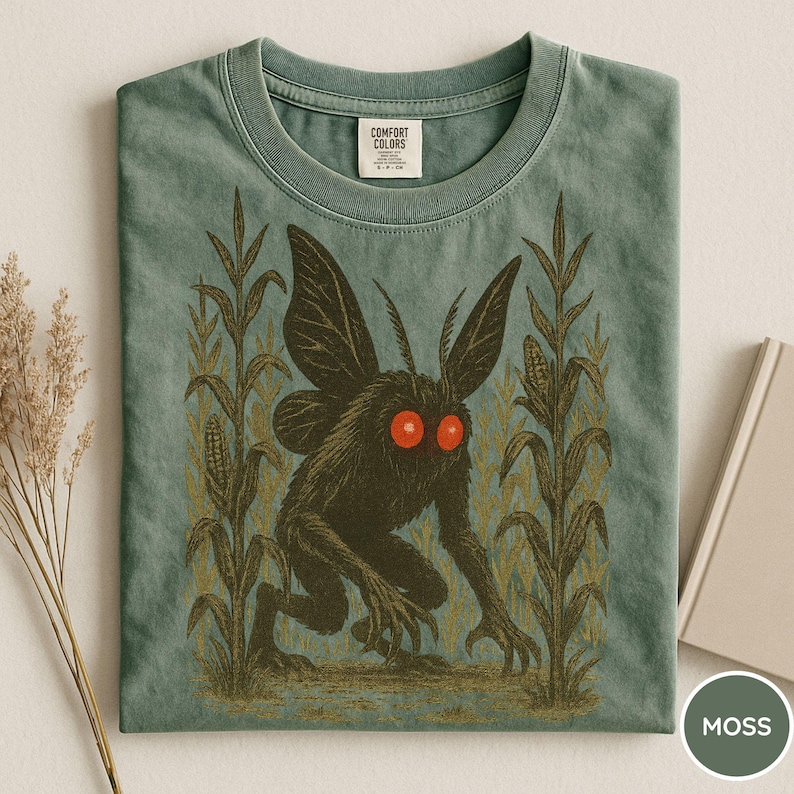 K&ouml;nnte beinhalten: Moosgr&uuml;nes T-Shirt mit einem schwarzen Wesen mit roten Augen und Fl&uuml;geln, umgeben von Maisstauden. Das T-Shirt hat einen Rundhalsausschnitt und ein Comfort Colors-Etikett.