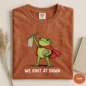 May include: A yam-colored t-shirt featuring a cartoon frog superhero with a red cape, holding knitting needles and yarn. The text "WE KNIT AT DAWN" is printed below the frog. The shirt has a crew neck and a Comfort Colors tag.