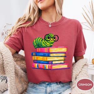 Pu&ograve; includere: Una t-shirt a maniche corte color cremisi con una stampa grafica di un bruco verde a righe che indossa occhiali tondi neri, appollaiato sopra una pila di cinque libri colorati nelle tonalit&agrave; del rosa, blu e giallo.