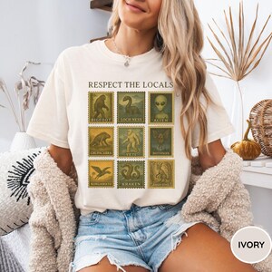 Pu&ograve; includere: T-shirt color avorio con un design a forma di francobollo con illustrazioni di criptidi, tra cui Bigfoot, il mostro di Loch Ness e Alien. Il testo "RESPECT THE LOCALS" &egrave; sopra i francobolli. La maglietta &egrave; girocollo e ha maniche corte.