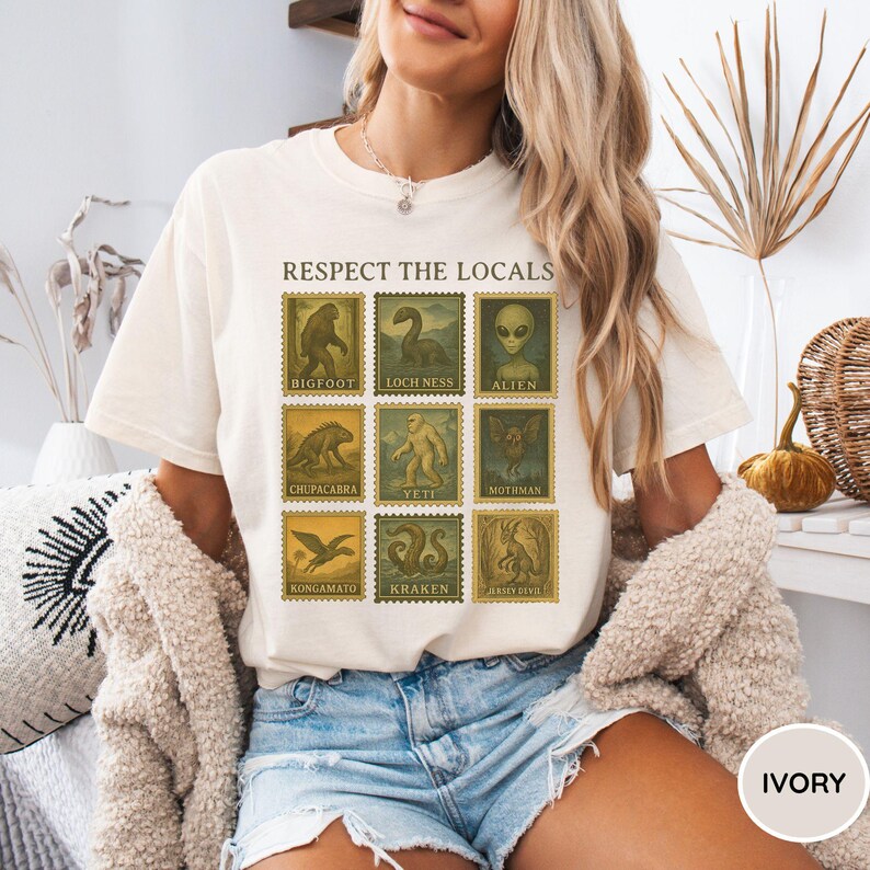 Pu&ograve; includere: T-shirt color avorio con un design a forma di francobollo con illustrazioni di criptidi, tra cui Bigfoot, il mostro di Loch Ness e Alien. Il testo "RESPECT THE LOCALS" &egrave; sopra i francobolli. La maglietta &egrave; girocollo e ha maniche corte.