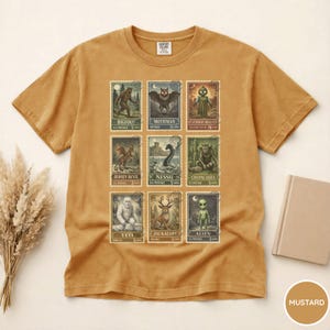 May include: Mustard-colored t-shirt featuring a collection of cryptid-themed postage stamp designs. The stamps depict creatures like Bigfoot, Mothman, and Nessie. The shirt has a classic crew neck and short sleeves, made of a soft, comfortable material.