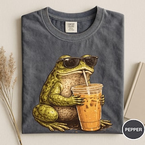 May include: Dark gray t-shirt featuring a cartoon frog wearing sunglasses and drinking iced coffee. The frog is green and tan, holding a clear plastic cup with a straw. The shirt has a Comfort Colors tag.
