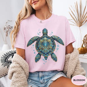 May include: A light pink short-sleeve t-shirt featuring a large, detailed graphic of a sea turtle with intricate blue and gold mandala-style patterns on its shell and flippers, surrounded by small blue bubbles on the front of the garment.