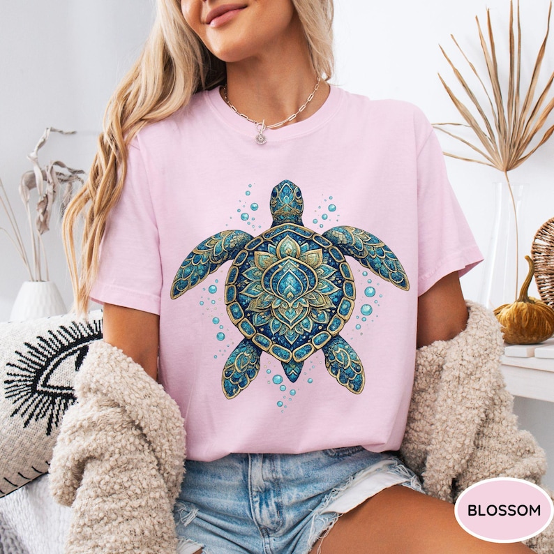 May include: A light pink short-sleeve t-shirt featuring a large, detailed graphic of a sea turtle with intricate blue and gold mandala-style patterns on its shell and flippers, surrounded by small blue bubbles on the front of the garment.