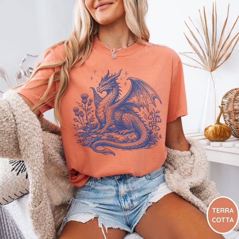 Peut inclure: T-shirt couleur terre cuite avec une illustration de dragon bleu. Le dragon est entour&eacute; de fleurs et d'&eacute;toiles bleues. Le t-shirt est &agrave; col rond et &agrave; manches courtes.