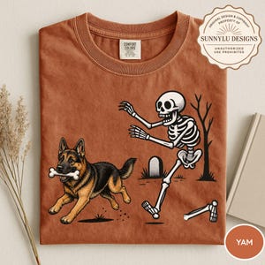 以下が含まれることがあります： 錆色のTシャツで、骨を口にくわえて走るジャーマンシェパードと、それを墓地で追いかける骸骨のグラフィックが描かれています。首元にはComfort Colorsのラベルがあり、SunnyLu Designsの透かしが入っています。
