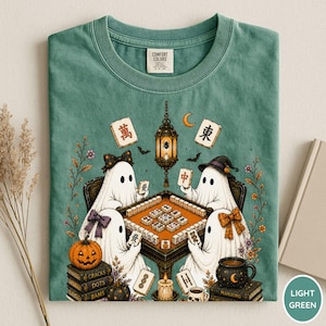 May include: A light green t-shirt featuring a graphic of four ghosts playing mahjong at a table. The design includes a hanging lantern, a pumpkin, stacks of books labeled Cracks, Dots, Bams, and Mahjong, and various floral accents.