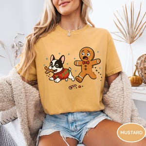 May include: Mustard-colored t-shirt featuring a cartoon dog in a Christmas sweater with a bone and a scared gingerbread man. The dog is black and white, and the gingerbread man is brown. The shirt is paired with denim shorts.