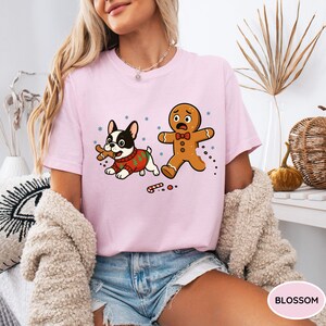 May include: A light pink t-shirt featuring a cartoon design of a French bulldog wearing a Christmas sweater and a gingerbread man. The dog is holding a bone, and the gingerbread man appears to be running away. Snowflakes and candy canes are also in the design.