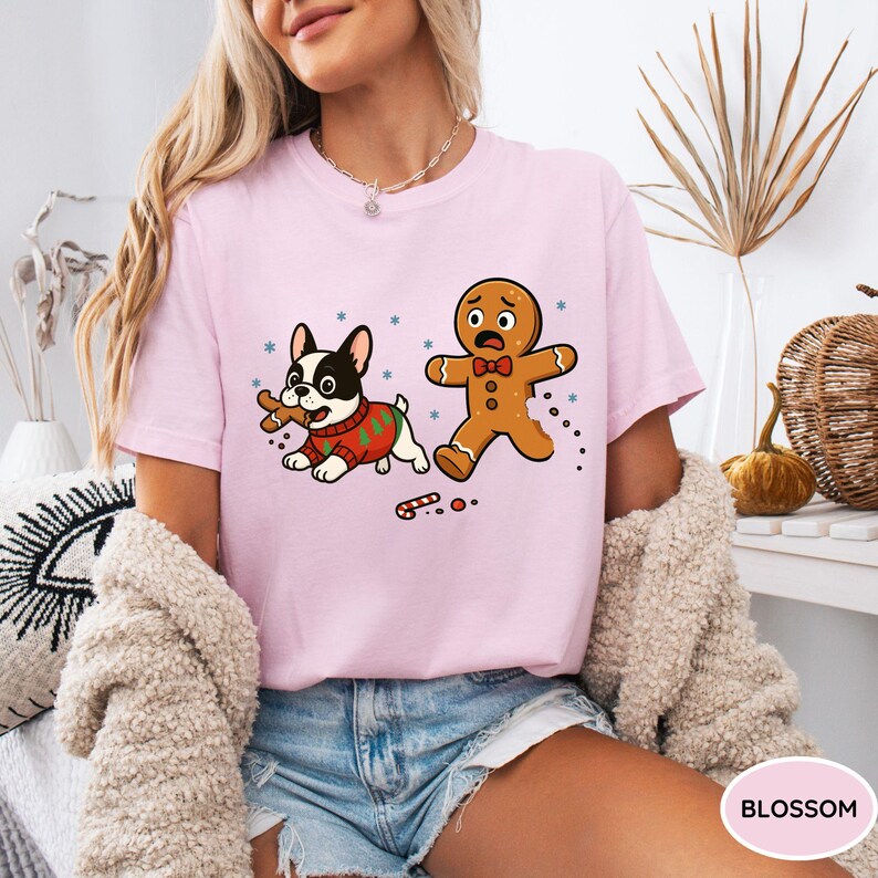 May include: A light pink t-shirt featuring a cartoon design of a French bulldog wearing a Christmas sweater and a gingerbread man. The dog is holding a bone, and the gingerbread man appears to be running away. Snowflakes and candy canes are also in the design.