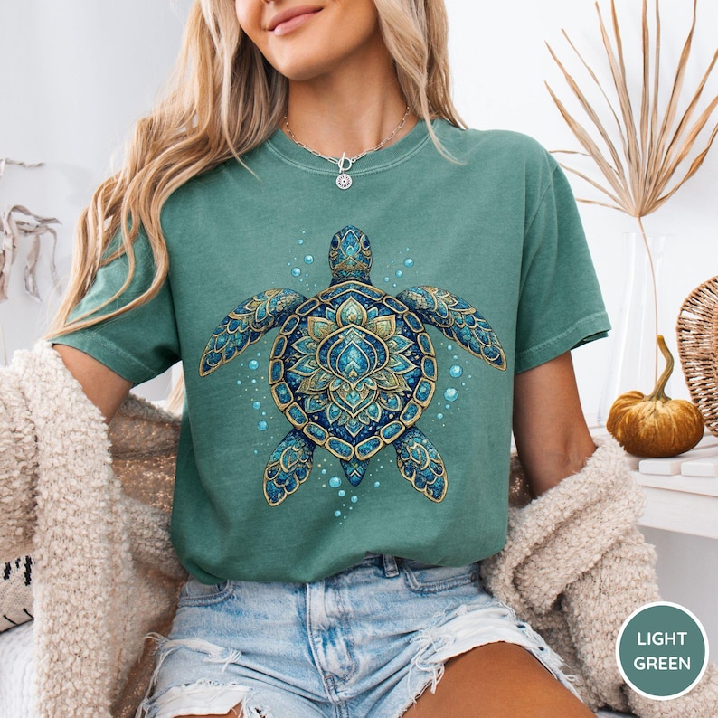 May include: A light green short-sleeve t-shirt featuring a large, detailed graphic of a sea turtle. The turtle is decorated with intricate blue and gold mandala-style patterns on its shell and flippers, surrounded by small light blue bubbles.