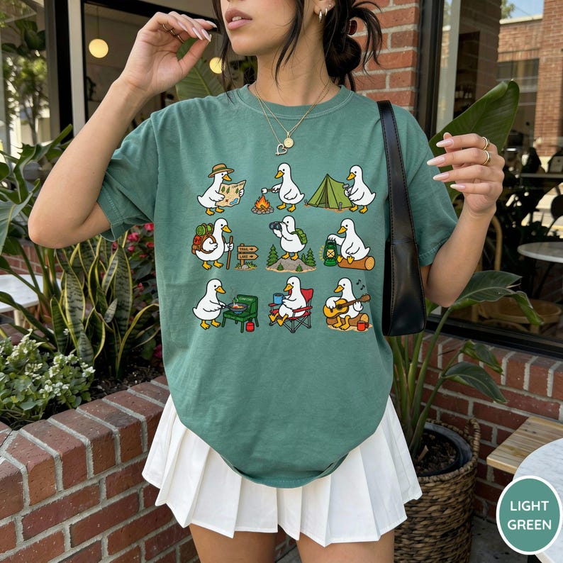 May include: A sage green t-shirt featuring a grid of nine cartoon ducks engaged in various camping activities, including hiking, roasting marshmallows, setting up a tent, using binoculars, holding a lantern, cooking, sitting in a chair, and playing guitar.