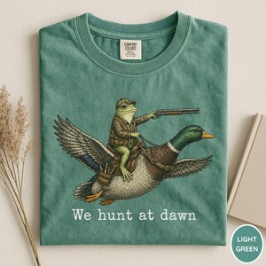 Puede incluir: Camiseta verde claro con una ilustraci&oacute;n caprichosa de una rana con atuendo de caza montada en un pato y sosteniendo una escopeta. El texto "We hunt at dawn" est&aacute; impreso debajo. Marca Comfort Colors.