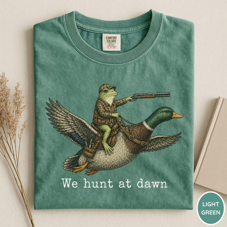 Puede incluir: Camiseta verde claro con una ilustraci&oacute;n caprichosa de una rana con atuendo de caza montada en un pato y sosteniendo una escopeta. El texto "We hunt at dawn" est&aacute; impreso debajo. Marca Comfort Colors.