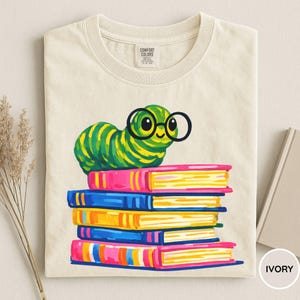 Pu&ograve; includere: Una t-shirt color avorio con la grafica di un bruco a strisce verdi e gialle che indossa occhiali tondi neri, appoggiato su una pila di cinque libri colorati nelle tonalit&agrave; del rosa, blu e giallo. L'etichetta interna riporta Comfort Colors.