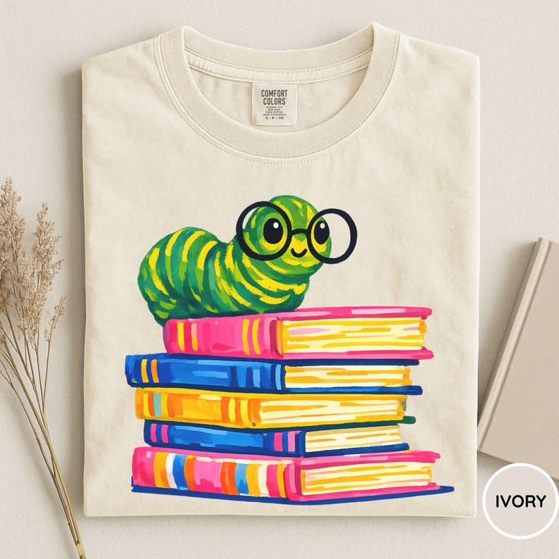 Pu&ograve; includere: Una t-shirt color avorio con la grafica di un bruco a strisce verdi e gialle che indossa occhiali tondi neri, appoggiato su una pila di cinque libri colorati nelle tonalit&agrave; del rosa, blu e giallo. L'etichetta interna riporta Comfort Colors.