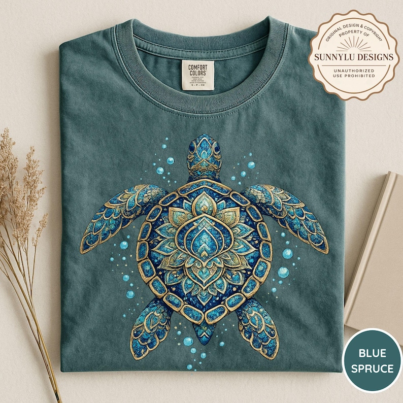 May include: A teal crewneck t-shirt featuring a central graphic of a sea turtle with an intricate blue and gold mandala-style shell pattern. The design includes small floating bubbles surrounding the turtle. The shirt has a Comfort Colors label.