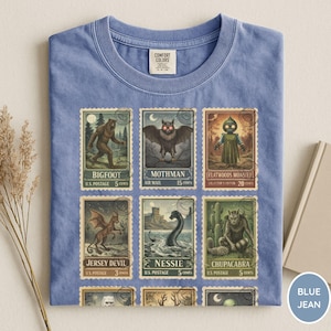 May include: Blue jean t-shirt featuring a collection of vintage-style postage stamps with cryptid illustrations, including Bigfoot, Mothman, and Nessie. The shirt has a crew neck and short sleeves. The Comfort Colors tag is visible.