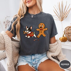 May include: Dark gray t-shirt featuring a cartoon dog in a Christmas sweater with a bone and a scared gingerbread man. The design includes snowflakes and candy canes, with the word "PEPPER" in a circle.