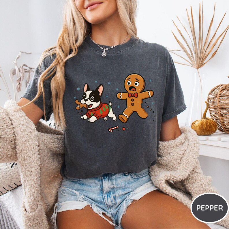 May include: Dark gray t-shirt featuring a cartoon dog in a Christmas sweater with a bone and a scared gingerbread man. The design includes snowflakes and candy canes, with the word "PEPPER" in a circle.