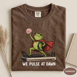 May include: A brown t-shirt featuring a green frog wearing a red cape, holding a pink ball labeled Pilates, and standing on a Pilates reformer. The text below the graphic reads WE PULSE AT DAWN. The shirt has a Comfort Colors brand neck label.