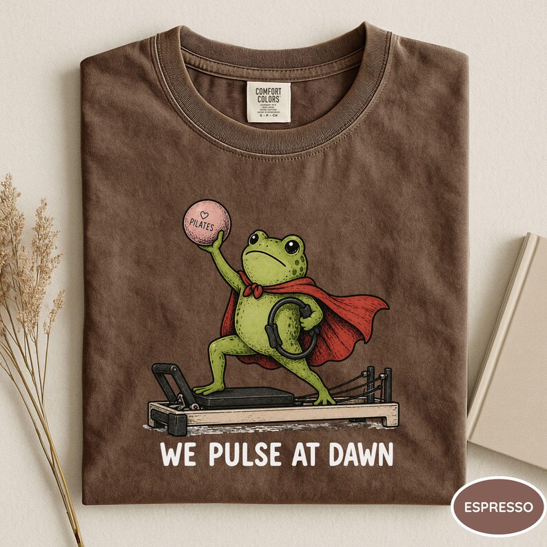 May include: A brown t-shirt featuring a green frog wearing a red cape, holding a pink ball labeled Pilates, and standing on a Pilates reformer. The text below the graphic reads WE PULSE AT DAWN. The shirt has a Comfort Colors brand neck label.