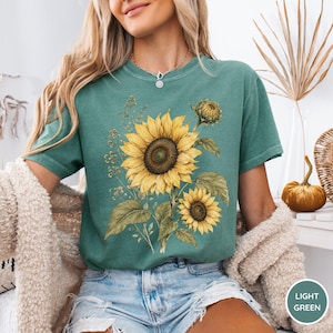 May include: A light green t-shirt featuring a watercolor sunflower design with yellow petals and green leaves. The shirt has a relaxed fit and short sleeves. The text "LIGHT GREEN" is in a white box in the lower right corner.