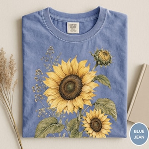 May include: A blue jean colored t-shirt featuring a watercolor illustration of sunflowers. The design includes a large, fully bloomed sunflower, a smaller one, and a sunflower bud, with green leaves and stems. The shirt has a crew neck and a tag that says "Comfort Colors".