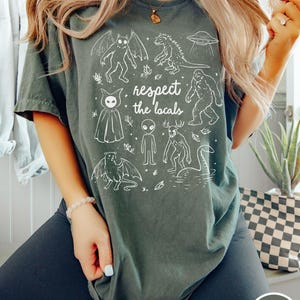 May include: Moss green t-shirt with a white graphic of various cryptids and the text "respect the locals". The design includes a bat-like creature, a Godzilla-like monster, a UFO, and Bigfoot. The shirt is a comfortable, casual style.