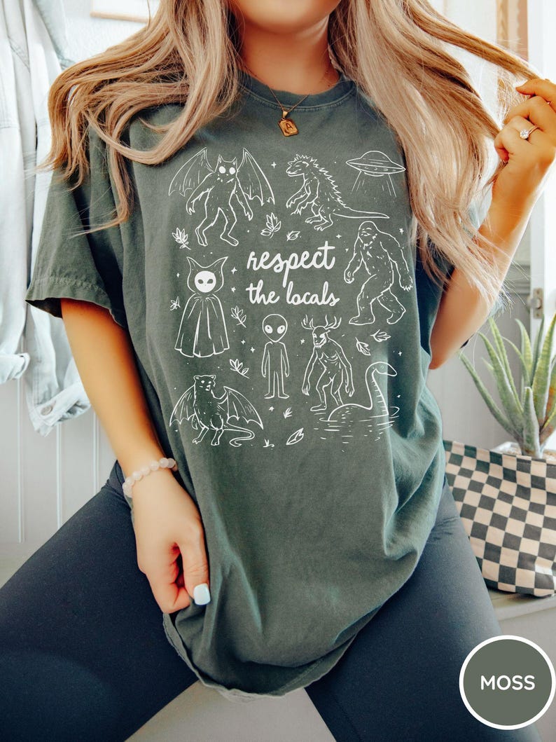 May include: Moss green t-shirt with a white graphic of various cryptids and the text "respect the locals". The design includes a bat-like creature, a Godzilla-like monster, a UFO, and Bigfoot. The shirt is a comfortable, casual style.