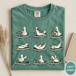 K&ouml;nnte beinhalten: Hellgr&uuml;nes T-Shirt mit einer verspielten Grafik von wei&szlig;en Enten, die auf Pilates-Reformern trainieren. Die Enten sind in verschiedenen Posen dargestellt, was dem Design eine skurrile Note verleiht. Das Shirt hat einen klassischen Rundhalsausschnitt.
