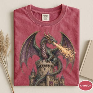 May include: A crimson short-sleeved t-shirt featuring a graphic of a green dragon perched atop a stone castle tower, breathing orange and yellow flames. The shirt has a crew neckline and a Comfort Colors brand label printed on the inside collar.